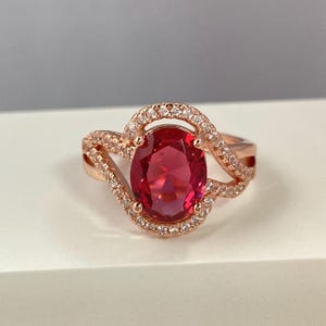 Peut inclure: Bague en or rose ornée d'une grande pierre rouge rubis de forme ovale. La pierre est entourée d'un halo de petites pierres claires. Le bandeau présente un design torsadé avec des pierres supplémentaires.