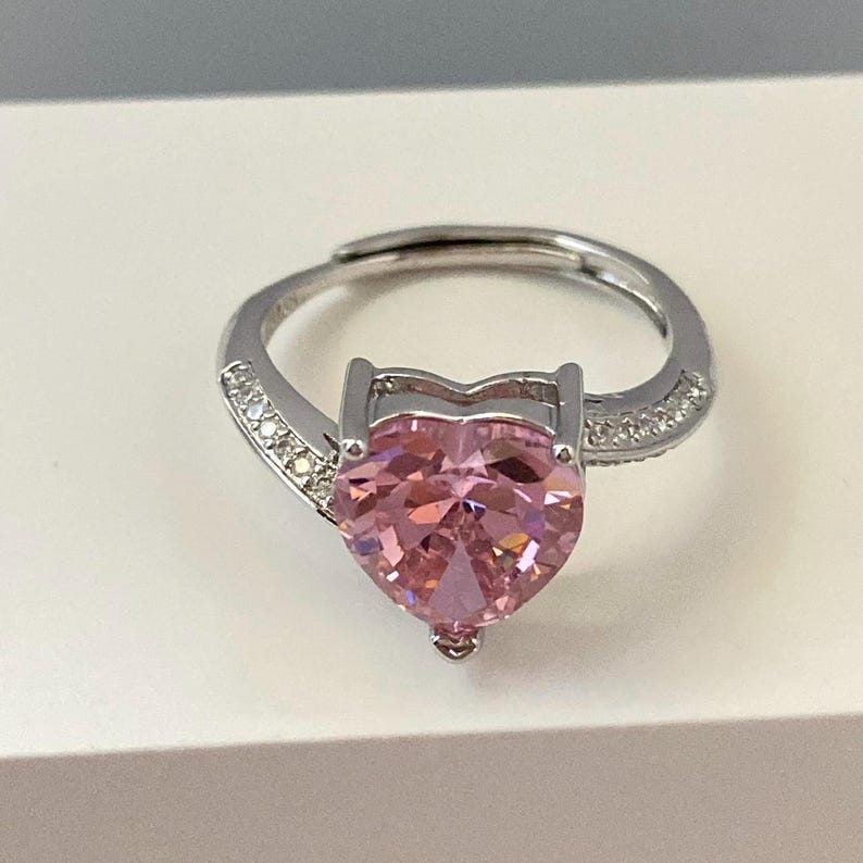 K&ouml;nnte beinhalten: Ein silberner Ring mit einem gro&szlig;en rosafarbenen herzf&ouml;rmigen Edelstein und kleineren klaren Edelsteinen auf der Ringschiene.