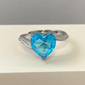 K&ouml;nnte beinhalten: Ein silberner Ring mit einem gro&szlig;en, herzf&ouml;rmigen blauen Edelstein und kleineren klaren Edelsteinen auf der Ringschiene.