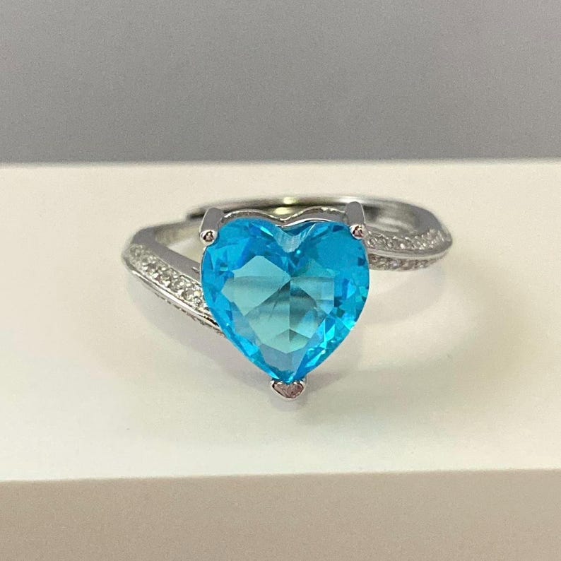 K&ouml;nnte beinhalten: Ein silberner Ring mit einem gro&szlig;en, herzf&ouml;rmigen blauen Edelstein und kleineren klaren Edelsteinen auf der Ringschiene.