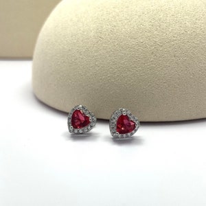 May include: A pair of silver heart-shaped earrings with a red gemstone in the center and a halo of smaller clear gemstones.