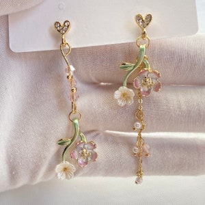 Puede incluir: Un par de pendientes colgantes de oro con un delicado diseño floral. Cada pendiente presenta un pequeño dije en forma de corazón en la parte superior, seguido de una cadena de cuentas rosadas y transparentes, y luego un tallo verde con dos flores blancas y rosadas. La parte inferior de cada pendiente tiene una cadena con pequeñas perlas blancas y cuentas rosadas.