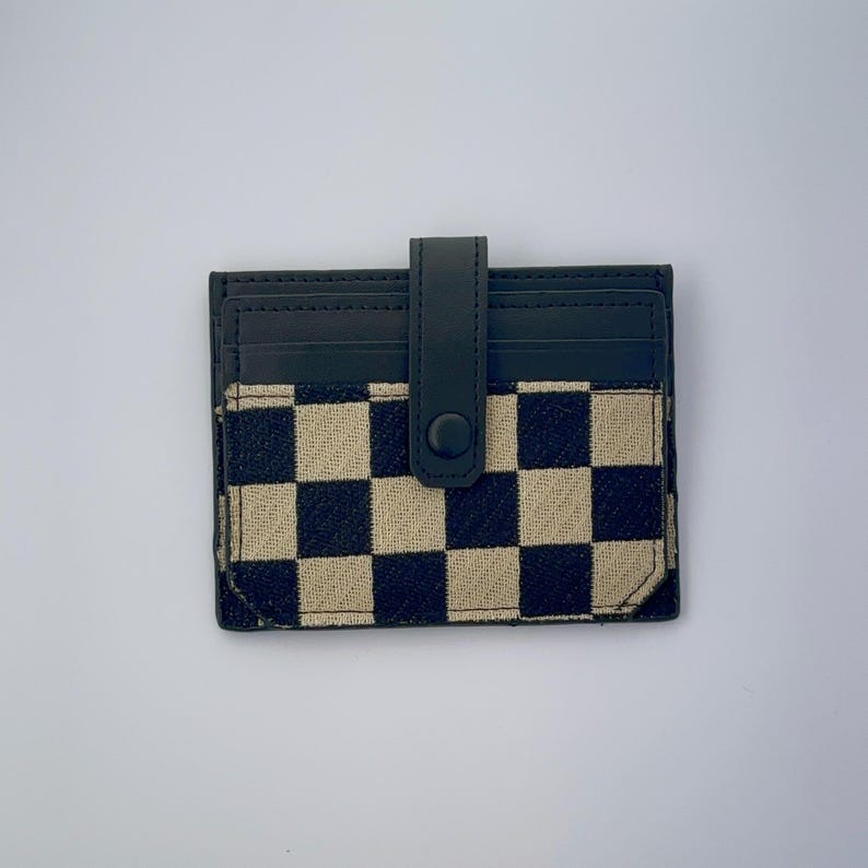 Puede incluir: Un porta tarjetas a cuadros blanco y negro con una solapa de cuero negro y cierre a presi&oacute;n.