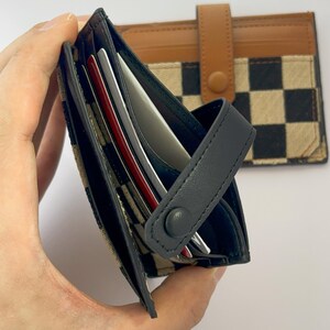 Puede incluir: Una cartera de cuero negro con un patr&oacute;n a cuadros y un cierre de solapa. La cartera tiene m&uacute;ltiples ranuras para tarjetas y un cierre a presi&oacute;n.
