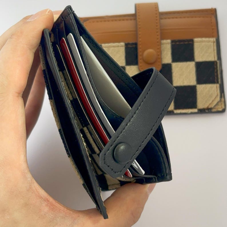 Puede incluir: Una cartera de cuero negro con un patr&oacute;n a cuadros y un cierre de solapa. La cartera tiene m&uacute;ltiples ranuras para tarjetas y un cierre a presi&oacute;n.