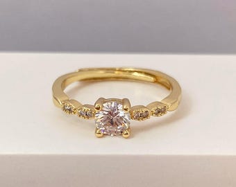 May include: A gold ring featuring a single, round, clear stone in the centre. The band is adorned with smaller, clear stones.