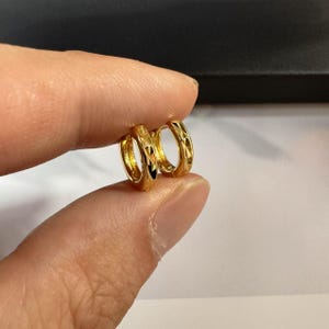 Puede incluir: Un par de pendientes de aro pequeños, de color dorado. Cada pendiente tiene un patrón geométrico de formas de diamante. Los pendientes se sostienen entre dos dedos, mostrando su tamaño y diseño. La joyería es brillante y reflectante.