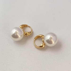 May include: A pair of gold-colored hoop earrings, each featuring a large, round, white pearl. The earrings are displayed on a white surface, showcasing their elegant design and reflective surfaces. The pearl earrings are a classic jewelry item.