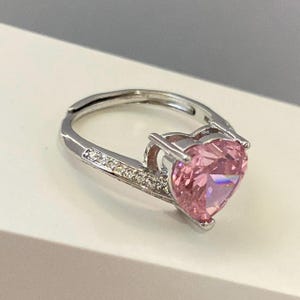 K&ouml;nnte beinhalten: Ein silberner Ring mit einem gro&szlig;en rosafarbenen herzf&ouml;rmigen Edelstein, der von kleineren klaren Edelsteinen umgeben ist.