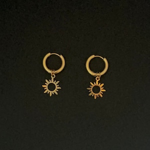 Puede incluir: Un par de pendientes de aro dorados con un pequeño encanto de sol colgando de cada aro.