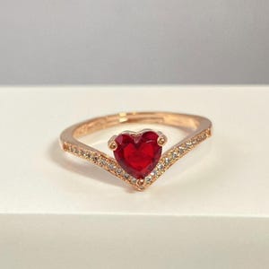 Puede incluir: Anillo de oro rosa con una piedra preciosa roja en forma de corazón. El anillo tiene una banda en forma de V con pequeñas piedras claras. La piedra central en forma de corazón está engastada en un delicado engaste de garras.