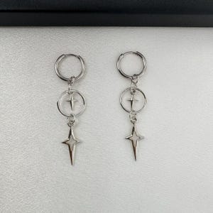 May include: A pair of silver hoop earrings with a star charm dangling from each hoop. The star charms have a hollow center.
