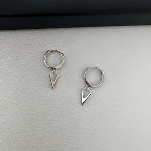 May include: A pair of silver hoop earrings with a small silver V charm hanging from each hoop.