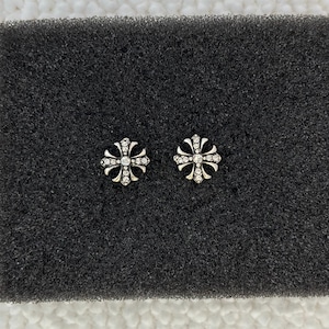 May include: A pair of silver-toned stud earrings shaped like ornate crosses. Each earring features a central clear stone surrounded by smaller clear stones embedded along the four arms of the cross, creating a sparkling, detailed metallic design.