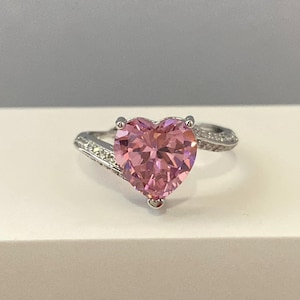K&ouml;nnte beinhalten: Ein silberner Ring mit einem gro&szlig;en rosafarbenen herzf&ouml;rmigen Edelstein und kleineren klaren Edelsteinen auf der Ringschiene.