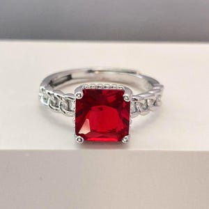 May include: A silver ring with a large, square, red gemstone set in the center. The band of the ring is a chain link design.