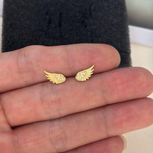 Puede incluir: Un par de pendientes de botón dorados con forma de alas de ángel. Cada ala presenta un diseño de plumas texturizado y está adornada con pequeñas piedras claras y brillantes incrustadas en la parte superior de la estructura.