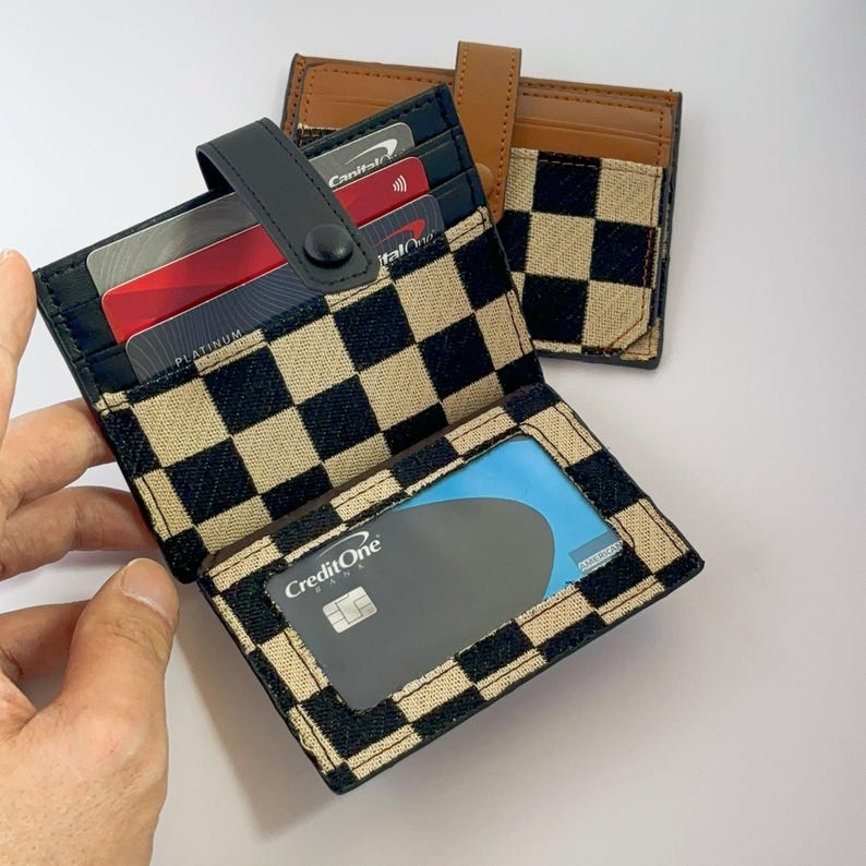 Puede incluir: Una cartera a cuadros negros y blancos con una solapa de cuero marr&oacute;n. La cartera tiene varias ranuras para tarjetas y una ventana de pl&aacute;stico transparente para mostrar una tarjeta de cr&eacute;dito. La cartera est&aacute; abierta y muestra una tarjeta CreditOne en el interior.