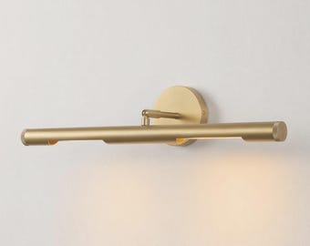 May include: A wall-mounted picture light with a cylindrical brass-toned metal bar and a circular mounting plate. The fixture features an adjustable arm connecting the horizontal tube to the base, emitting a soft glow from the underside.