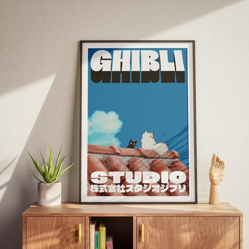 May include: A framed poster with a blue background and white text that reads "Studio Ghibli" in a bold, stylized font. The poster features a black cat and two white cats sitting on a red tiled roof.