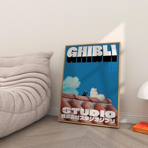May include: A framed poster with a blue background and the text "Studio Ghibli" in white and black. The text "株式会社スタジオジブリ" is in Japanese below the Studio Ghibli text. Two cats, one black and one white, are sitting on a red tiled roof.