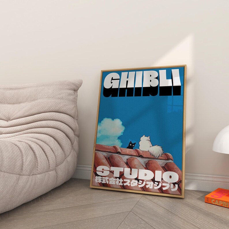 May include: A framed poster with a blue background and the text "Studio Ghibli" in white and black. The text "株式会社スタジオジブリ" is in Japanese below the Studio Ghibli text. Two cats, one black and one white, are sitting on a red tiled roof.