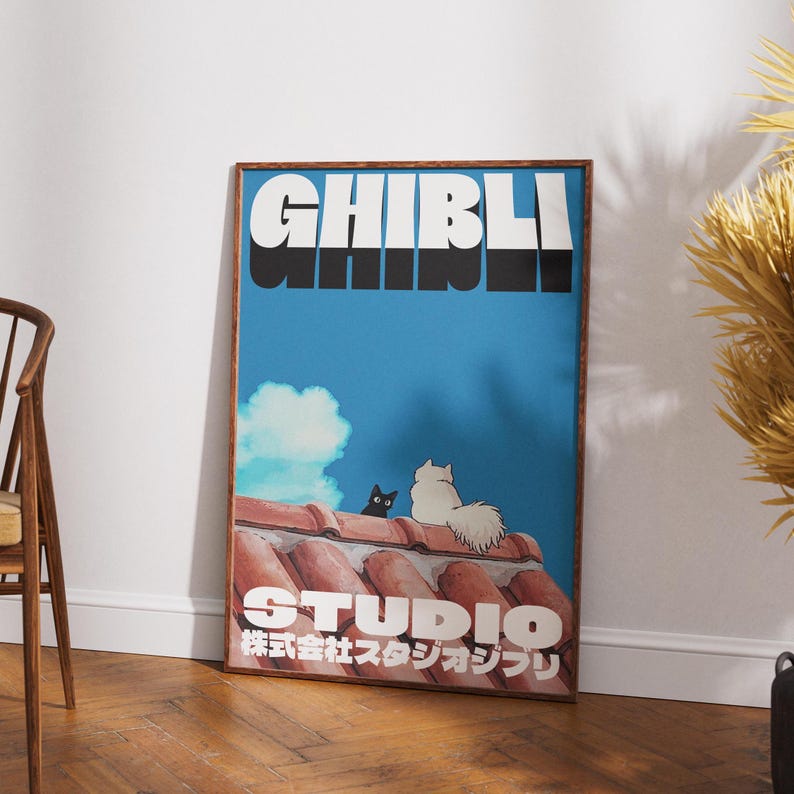 May include: A framed poster with a blue background featuring the text "Studio Ghibli" in white and black letters. The poster also features two cats, one black and one white, sitting on a red tiled roof.