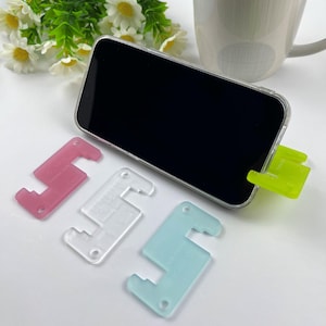 May include: A smartphone in a clear case is supported by a neon green stand. Three S-shaped phone stands in pink, clear, and blue are in front of the phone. Daisies and a mug are in the background.