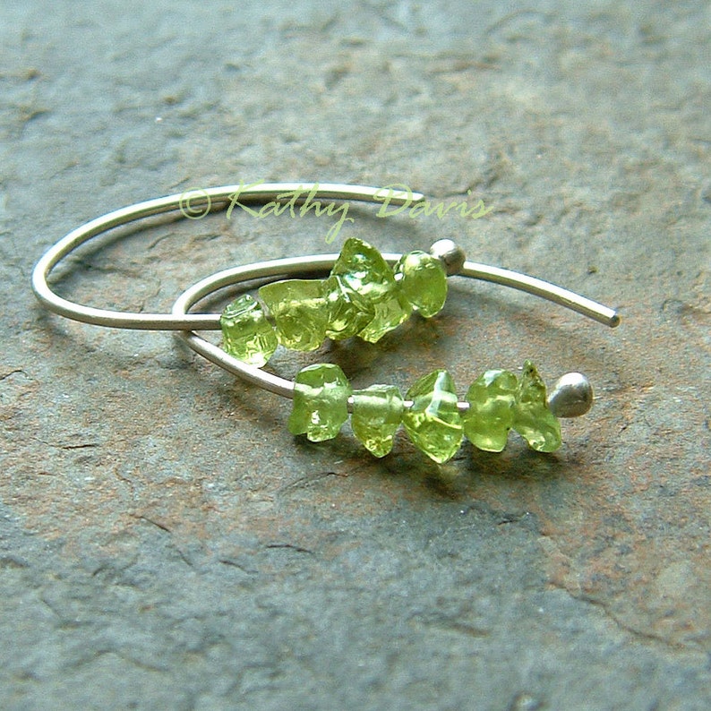 May include: Silver-tone earrings with a curved design, each featuring a row of small, light green peridot gemstones. The earrings are presented on a textured, grey surface.