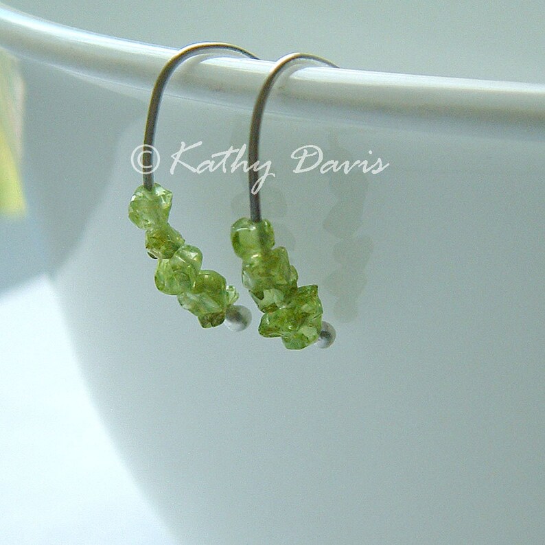 May include: Hoop earrings featuring clusters of light green peridot gemstones. The earrings have silver-coloured metal wires and small silver beads. The image includes the text "Kathy Davis".