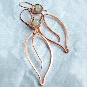 Puede incluir: Un par de pendientes de alambre de cobre con una piedra preciosa verde claro en cada uno. Los pendientes tienen forma de hojas y un dise&ntilde;o delicado y org&aacute;nico.