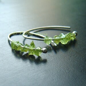 May include: A pair of silver hoop earrings, each featuring a line of small, irregularly shaped, light green peridot gemstones. The earrings have a simple, minimalist design, with a small silver bead at the end of each hoop.