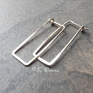 May include: A pair of gold-colored rectangular hoop earrings. The earrings have a simple, geometric design with a rectangular shape. The earrings are displayed on a gray surface. The earrings are lightweight and suitable for everyday wear.