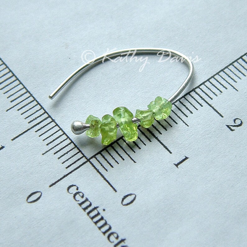 May include: A silver earring with a curved hook. It has a row of small, light green peridot gemstones. The gemstones are irregularly shaped, adding a natural element. The earring is shown next to a ruler marked in centimetres.