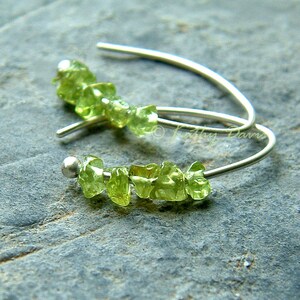 May include: Silver earrings with a row of small, light green peridot stones. The earrings have a curved design with a small silver ball at the end of each row of stones. The peridot stones are a vibrant, natural green colour.