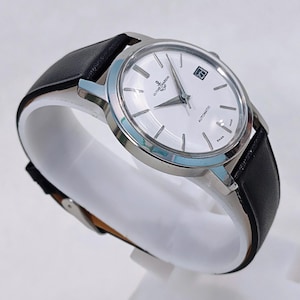 May include: A silver-toned Ulysse Nardin automatic wristwatch with a white face and black leather band. The watch has a date display and is marked "Swiss Made". The watch is displayed on a white stand.