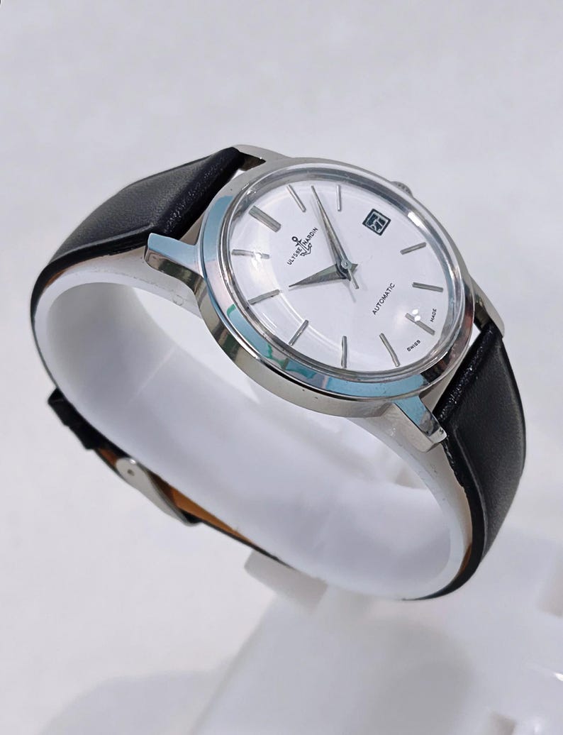 May include: A silver-toned Ulysse Nardin automatic wristwatch with a white face and black leather band. The watch has a date display and is marked "Swiss Made". The watch is displayed on a white stand.