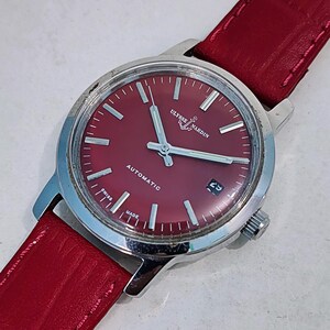 May include: A vintage Ulysse Nardin automatic wristwatch with a red dial and a red leather strap. The watch has silver hands and hour markers, a date window at the 3 o'clock position, and the words "Ulysse Nardin", "Automatic", "Swiss Made" are visible.