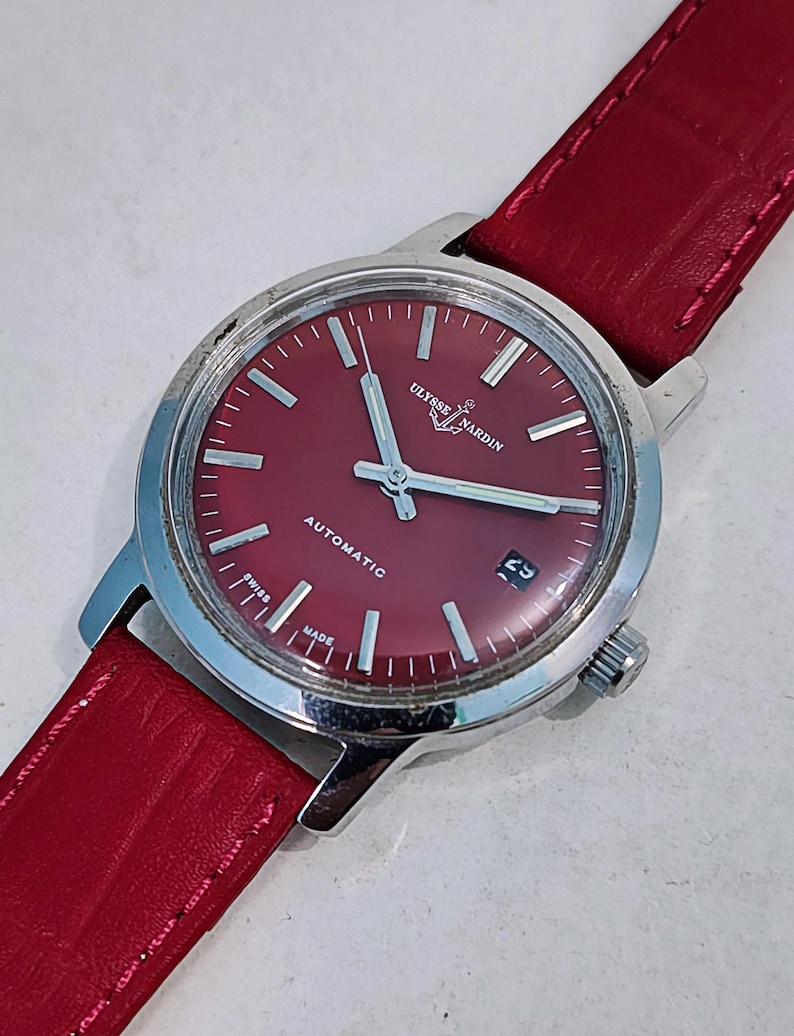 May include: A vintage Ulysse Nardin automatic wristwatch with a red dial and a red leather strap. The watch has silver hands and hour markers, a date window at the 3 o'clock position, and the words "Ulysse Nardin", "Automatic", "Swiss Made" are visible.
