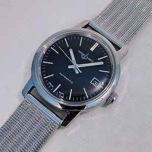 May include: A silver-toned Ulysse Nardin automatic wristwatch with a black dial and a date display. The watch features silver hands and markers, and a stainless steel mesh band. The watch is marked "Swiss Made".