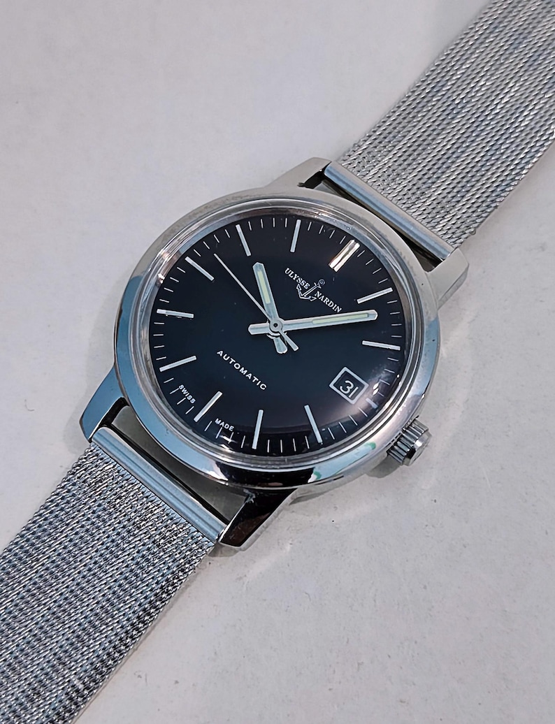 May include: A silver-toned Ulysse Nardin automatic wristwatch with a black dial and a date display. The watch features silver hands and markers, and a stainless steel mesh band. The watch is marked "Swiss Made".