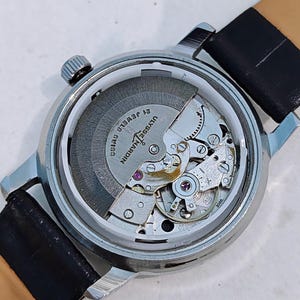 K&ouml;nnte beinhalten: Nahaufnahme einer silberfarbenen Armbanduhr mit transparentem Geh&auml;useboden, die das Innenleben zeigt. Die Uhr hat ein schwarzes Lederarmband und den Text "ULYSSE NARDIN SWISS 21 JEWELS".