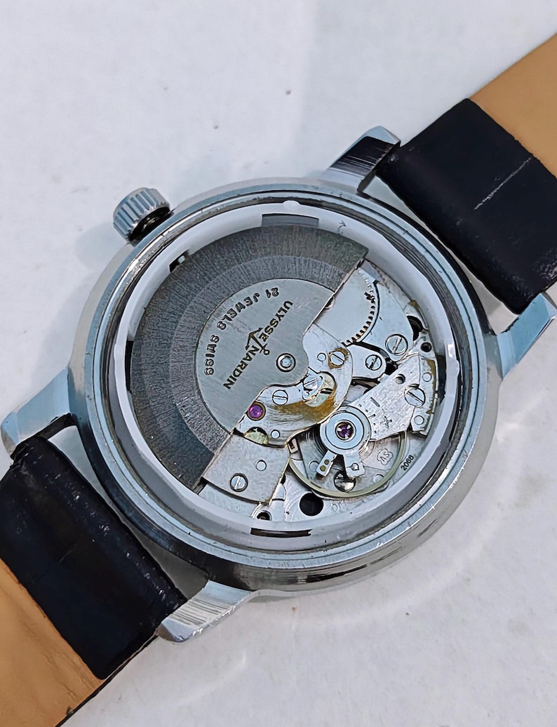 K&ouml;nnte beinhalten: Nahaufnahme einer silberfarbenen Armbanduhr mit transparentem Geh&auml;useboden, die das Innenleben zeigt. Die Uhr hat ein schwarzes Lederarmband und den Text "ULYSSE NARDIN SWISS 21 JEWELS".