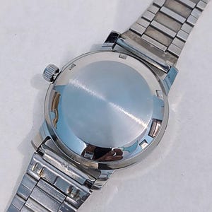 May include: A vintage wristwatch with a silver-toned case and a matching stainless steel band. The watch's back is polished and reflective, with a small crown on the side. The band is composed of rectangular links.