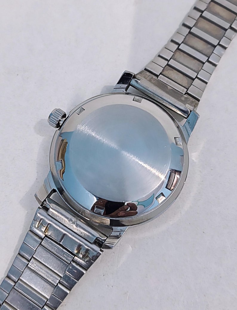 May include: A vintage wristwatch with a silver-toned case and a matching stainless steel band. The watch's back is polished and reflective, with a small crown on the side. The band is composed of rectangular links.