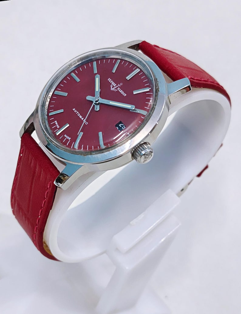 May include: A stainless steel wristwatch with a burgundy dial and a red leather strap. The watch face features silver-tone hands and markers, with the text "ULYSSE NARDIN" and "AUTOMATIC". The watch is displayed on a white stand.