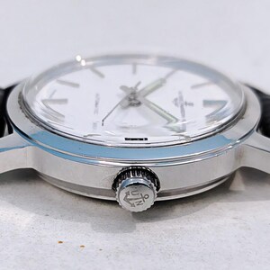 K&ouml;nnte beinhalten: Eine silberne Armbanduhr mit wei&szlig;em Zifferblatt und schwarzem Lederarmband. Die Uhr hat ein klares Glas und eine silberne Krone mit Logo. Die Zeiger der Uhr sind silber und gr&uuml;n. Die Uhr hat ein klassisches Design.