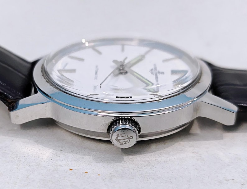 K&ouml;nnte beinhalten: Eine silberne Armbanduhr mit wei&szlig;em Zifferblatt und schwarzem Lederarmband. Die Uhr hat ein klares Glas und eine silberne Krone mit Logo. Die Zeiger der Uhr sind silber und gr&uuml;n. Die Uhr hat ein klassisches Design.