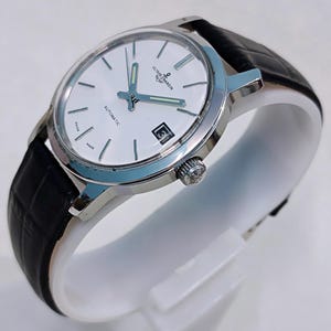 K&ouml;nnte beinhalten: Eine silberfarbene Armbanduhr mit wei&szlig;em Zifferblatt, schwarzem Lederarmband und Datumsanzeige auf 3 Uhr. Die Uhr tr&auml;gt die Aufschrift "ULYSSE NARDIN" und "AUTOMATIC" auf dem Zifferblatt. Die Uhr wird auf einem wei&szlig;en St&auml;nder pr&auml;sentiert.