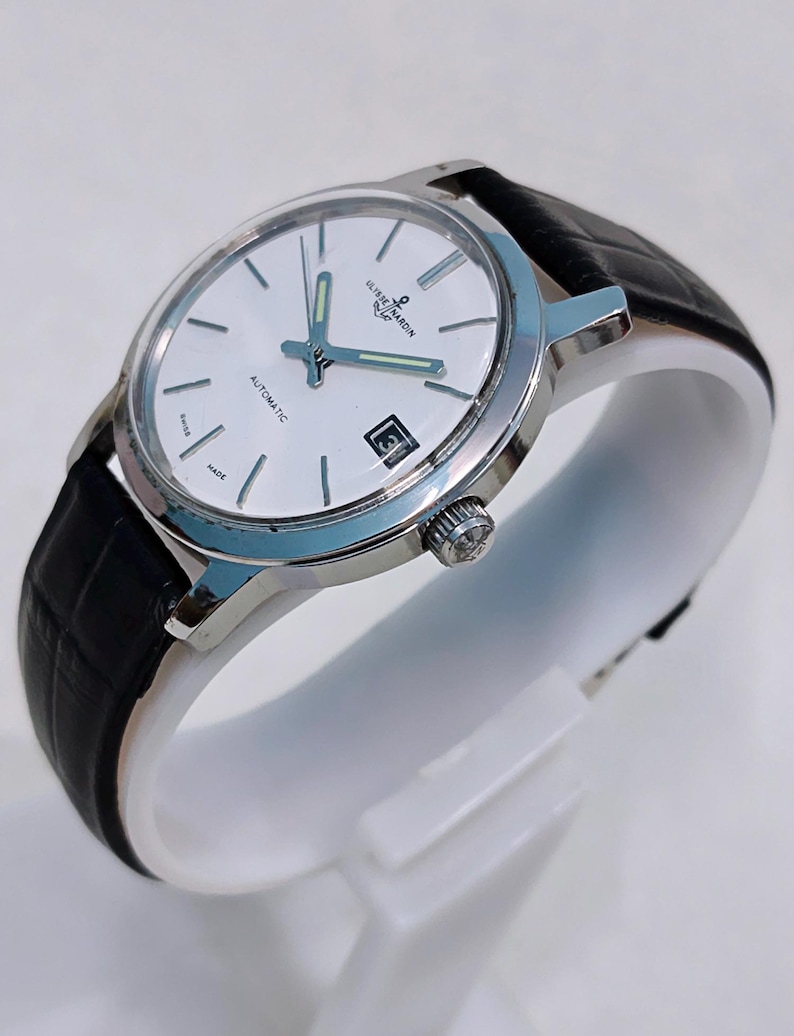 K&ouml;nnte beinhalten: Eine silberfarbene Armbanduhr mit wei&szlig;em Zifferblatt, schwarzem Lederarmband und Datumsanzeige auf 3 Uhr. Die Uhr tr&auml;gt die Aufschrift "ULYSSE NARDIN" und "AUTOMATIC" auf dem Zifferblatt. Die Uhr wird auf einem wei&szlig;en St&auml;nder pr&auml;sentiert.